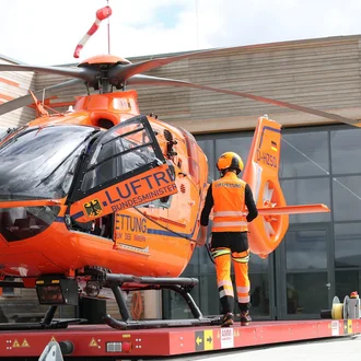 Luftretter vor orangefarbenem ADAC-Rettungshubschrauber beim Einsatzstart – Luftrettungsteam des Bundesministeriums des Innern auf Hubschrauberlandeplattform vor moderner Wache