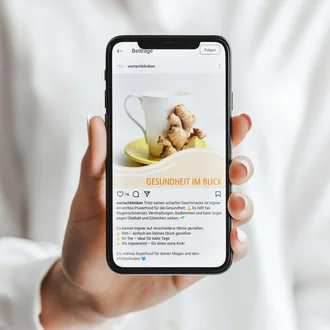 Hand hält ein Smartphone mit einem Instagram-Post der Wertachkliniken zum Thema Ingwer. Der Beitrag trägt den Titel „GESUNDHEIT IM BLICK“ und zeigt eine weiße Tasse auf einem gelben Teller mit frischem Ingwer. Der Text hebt die gesundheitlichen Vorteile von Ingwer hervor und gibt Tipps zur Einnahme – roh, als Tee oder in Aufgussform.