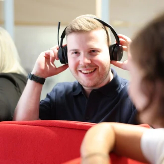 Mitarbeiter bei Denkströme setzt ein Headset auf und lächelt; im Hintergrund sitzen Kolleginnen an einer roten Sitznische.