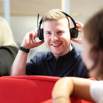 Mitarbeiter bei Denkströme setzt ein Headset auf und lächelt; im Hintergrund sitzen Kolleginnen an einer roten Sitznische.