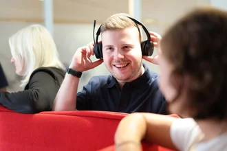 Mitarbeiter bei Denkströme setzt ein Headset auf und lächelt; im Hintergrund sitzen Kolleginnen an einer roten Sitznische.