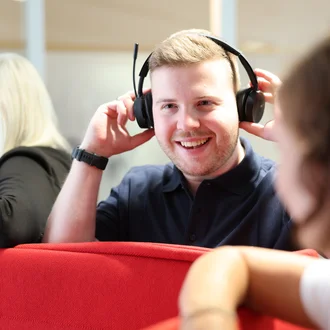 Mitarbeiter bei Denkströme setzt ein Headset auf und lächelt; im Hintergrund sitzen Kolleginnen an einer roten Sitznische.