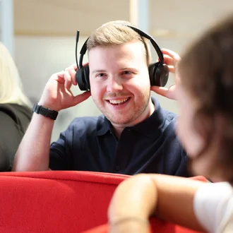 Mitarbeiter bei Denkströme setzt ein Headset auf und lächelt; im Hintergrund sitzen Kolleginnen an einer roten Sitznische.