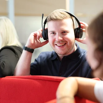 Mitarbeiter bei Denkströme setzt ein Headset auf und lächelt; im Hintergrund sitzen Kolleginnen an einer roten Sitznische.