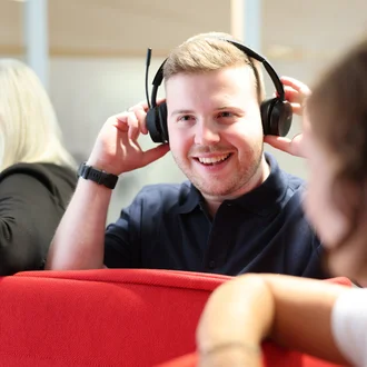 Mitarbeiter bei Denkströme setzt ein Headset auf und lächelt; im Hintergrund sitzen Kolleginnen an einer roten Sitznische.