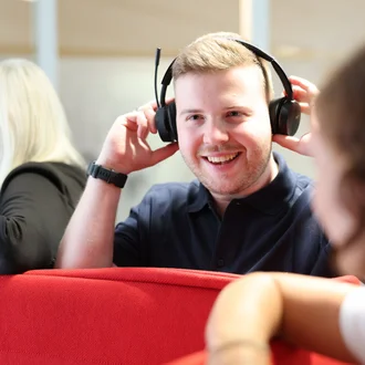 Mitarbeiter bei Denkströme setzt ein Headset auf und lächelt; im Hintergrund sitzen Kolleginnen an einer roten Sitznische.