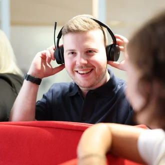 Mitarbeiter bei Denkströme setzt ein Headset auf und lächelt; im Hintergrund sitzen Kolleginnen an einer roten Sitznische.