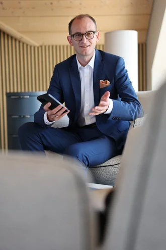 Fabian Schmid, leitender Geschäftsführer von Denkströme, sitzt in einem Lounge-Bereich, hält ein Smartphone in der Hand und spricht gestikulierend.