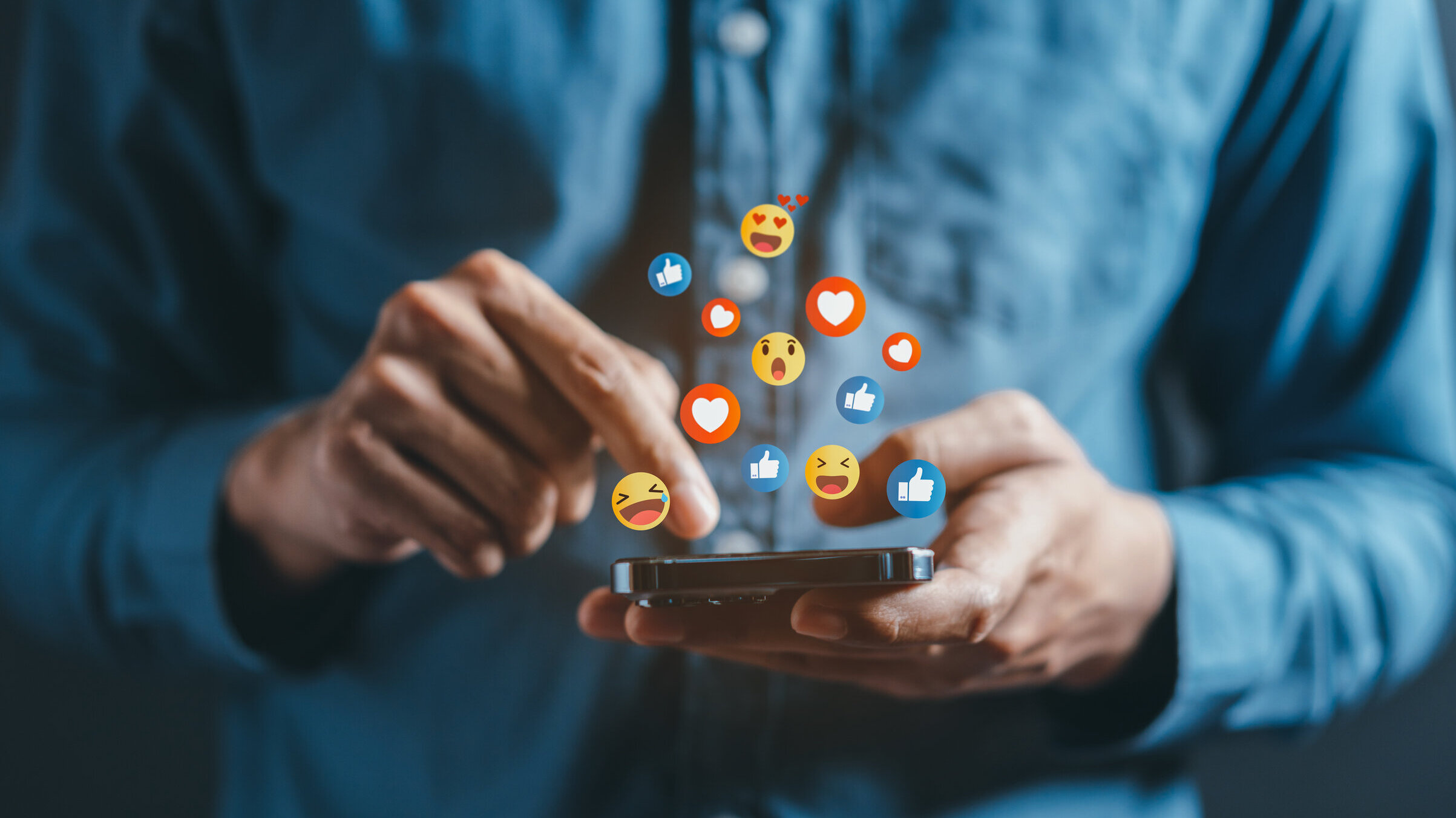 Person tippt auf Smartphone, während bunte Social-Media-Emojis wie Likes und Herzen darüber schweben