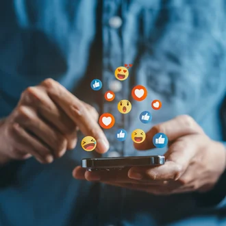 Person tippt auf Smartphone, während bunte Social-Media-Emojis wie Likes und Herzen darüber schweben