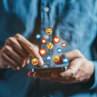 Person tippt auf Smartphone, während bunte Social-Media-Emojis wie Likes und Herzen darüber schweben