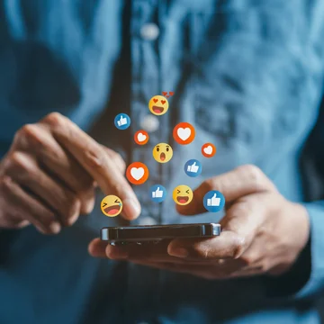 Person tippt auf Smartphone, während bunte Social-Media-Emojis wie Likes und Herzen darüber schweben