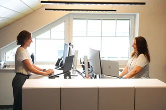 Zwei Mitarbeitende im Büro stehen an einem höhenverstellbaren Doppel-Schreibtisch und arbeiten am Computer; heller Raum mit großen Dachfenstern.