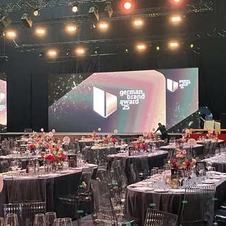 Der festlich dekorierte Veranstaltungssaal des German Brand Award 2025 kurz vor Beginn der Preisverleihung: Rund gedeckte Tische mit frischen Blumen, nummerierten Plätzen und stilvoller Beleuchtung schaffen eine exklusive Atmosphäre für die Ehrung herausragender Markenprojekte.