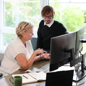 Projektsteuerung bei Denkströme: Zwei Mitarbeitende besprechen am Schreibtisch Inhalte am Monitor; Notizbuch, Tastatur und Kaffeetasse liegen bereit.