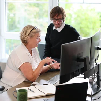 Projektsteuerung bei Denkströme: Zwei Mitarbeitende besprechen am Schreibtisch Inhalte am Monitor; Notizbuch, Tastatur und Kaffeetasse liegen bereit.