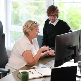 Projektsteuerung bei Denkströme: Zwei Mitarbeitende besprechen am Schreibtisch Inhalte am Monitor; Notizbuch, Tastatur und Kaffeetasse liegen bereit.