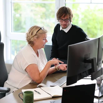 Projektsteuerung bei Denkströme: Zwei Mitarbeitende besprechen am Schreibtisch Inhalte am Monitor; Notizbuch, Tastatur und Kaffeetasse liegen bereit.
