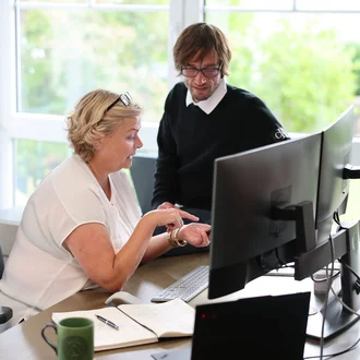 Projektsteuerung bei Denkströme: Zwei Mitarbeitende besprechen am Schreibtisch Inhalte am Monitor; Notizbuch, Tastatur und Kaffeetasse liegen bereit.