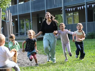 Erzieherin läuft mit vier lachenden Kindern über das Außengelände einer Kindertagesstätte