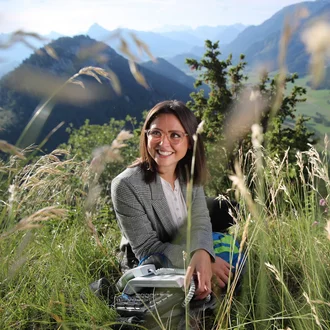 Lächelnde Verwaltungsmitarbeiterin mit Telefonanlage im Grünen vor Alpenpanorama – authentisches Bildmotiv für moderne Büroarbeit in naturnaher Umgebung bei der Fachklinik Allgäu.