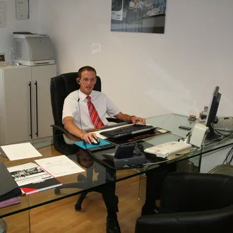 Büroaufnahme aus dem Jahr 2006: junger Mann mit Headset sitzt im weißen Hemd und roter Krawatte an einem Glastisch mit Röhrenmonitor und Telefonanlage