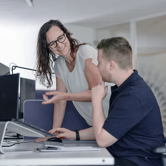 Zwei Mitarbeitende aus der Redaktion von Denkströme arbeiten am Schreibtisch: Eine Person zeigt auf den Monitor, die andere sitzt am Computer und hört zu.