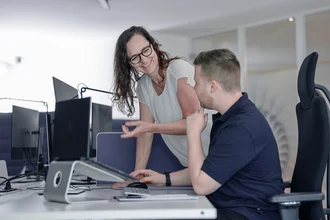 Zwei Mitarbeitende aus der Redaktion von Denkströme arbeiten am Schreibtisch: Eine Person zeigt auf den Monitor, die andere sitzt am Computer und hört zu.