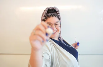 Mitarbeiterin aus der Grafikabteilung von Denkströme steht vor einem Whiteboard, lächelt in die Kamera und hält einen Whiteboard-Marker in der Hand.