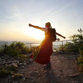 Mitarbeiterin im Servicebereich der Fachklinik Allgäu tanzt in Allgäuer Tracht mit Rucksack vor Sonnenuntergang – symbolstarkes Motiv der Arbeitgeberkampagne für Kulturverbundenheit und Lebensfreude am Arbeitsplatz.