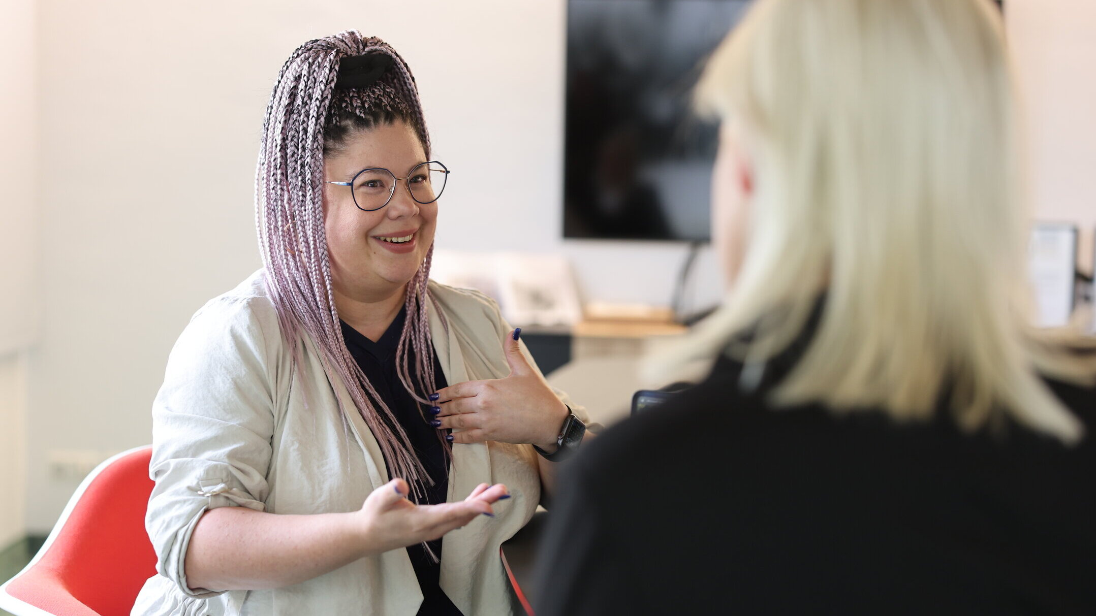 Denkströme Mitarbeiterin mit geflochtenen Haaren spricht gestikulierend mit einer Kollegin im Büro – Gesprächssituation für Social Media Recruiting und Employer Branding.