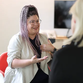 Denkströme Mitarbeiterin mit geflochtenen Haaren spricht gestikulierend mit einer Kollegin im Büro – Gesprächssituation für Social Media Recruiting und Employer Branding.