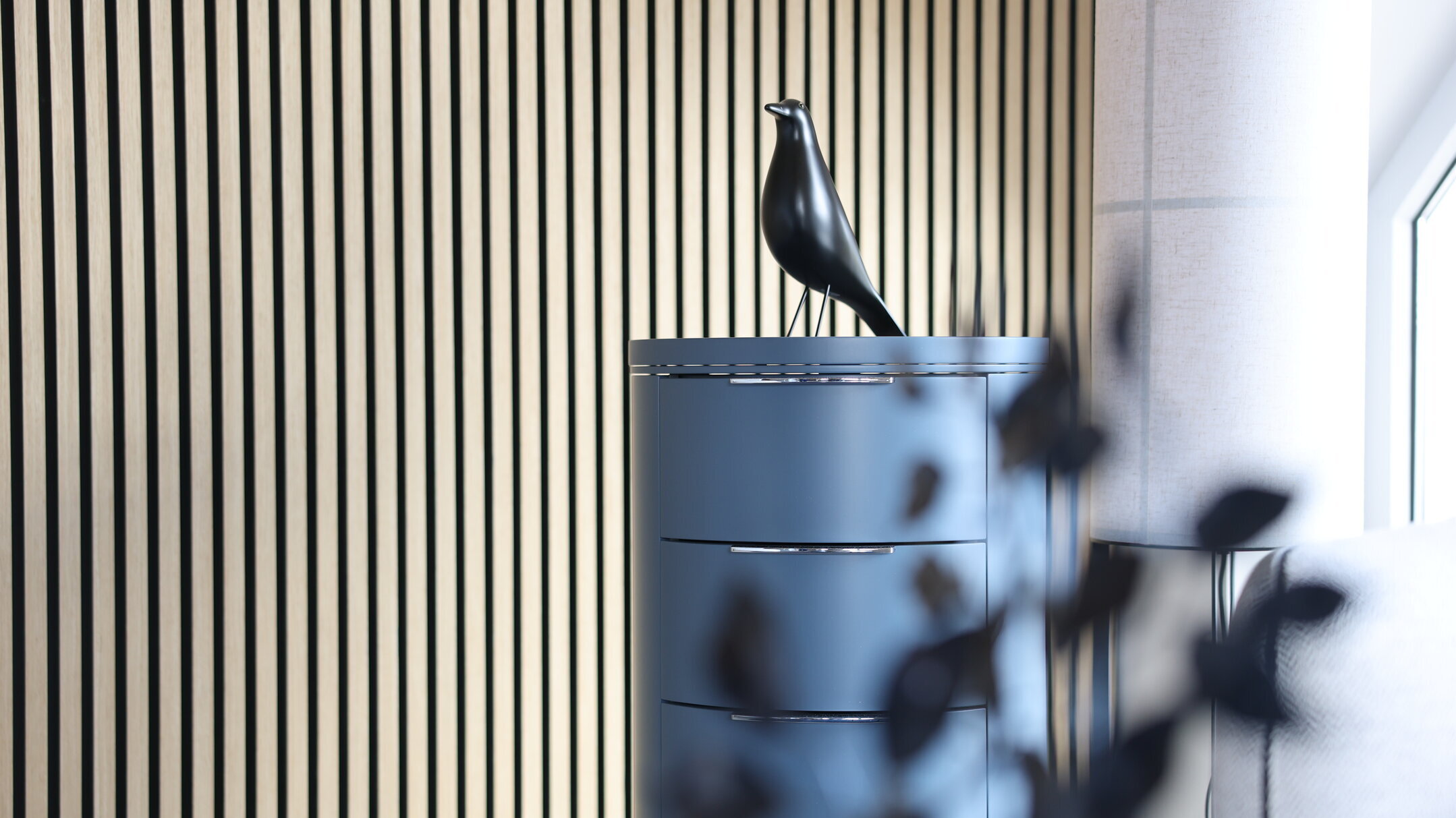 Detailaufnahme im Denkströme-Büro: Holzlamellenwand mit blauem Sideboard und schwarzer Vogelfigur als Deko, rechts ein Fenster mit Vorhang.