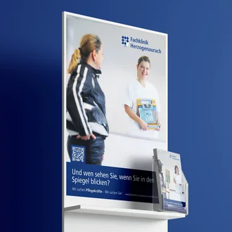 Recruiting-Display der Fachklinik Herzogenaurach mit Plakatmotiv und Broschürenhalter vor blauem Hintergrund zur Ansprache potenzieller Pflegekräfte.