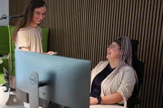 Zwei Mitarbeitende aus der Grafikabteilung von Denkströme lachen im Büro an einem Arbeitsplatz mit großem Monitor; eine Person sitzt am Schreibtisch, die andere steht daneben.