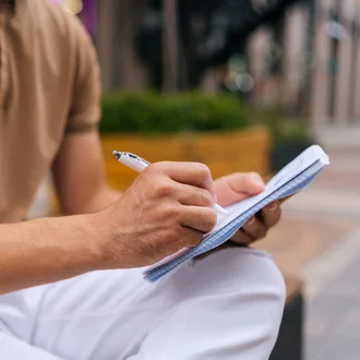 Person in beiger Kleidung schreibt mit einem Stift in ein kariertes Notizbuch im Freien