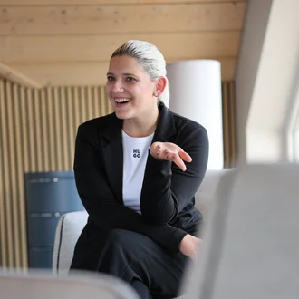 Teresa Schmid (Member of Management Board) sitzt in einem Besprechungsraum auf einem Sessel, lächelt und gestikuliert während eines Gesprächs.