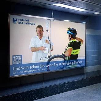 Leuchtendes Plakat der Fachklinik Bad Heilbrunn in U-Bahnstation mit Slogan zur Pflegekräftegewinnung, zeigt Pfleger und Radfahrer im Dialogmotiv.