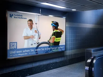 Leuchtendes Plakat der Fachklinik Bad Heilbrunn in U-Bahnstation mit Slogan zur Pflegekräftegewinnung, zeigt Pfleger und Radfahrer im Dialogmotiv.