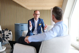 Beratungsgespräch bei Denkströme: Ein Manager im Anzug erklärt gestikulierend etwas in einem Lounge-Bereich, gegenüber sitzt ein Gesprächspartner.