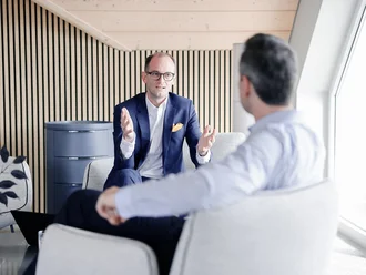Beratungsgespräch bei Denkströme: Ein Manager im Anzug erklärt gestikulierend etwas in einem Lounge-Bereich, gegenüber sitzt ein Gesprächspartner.