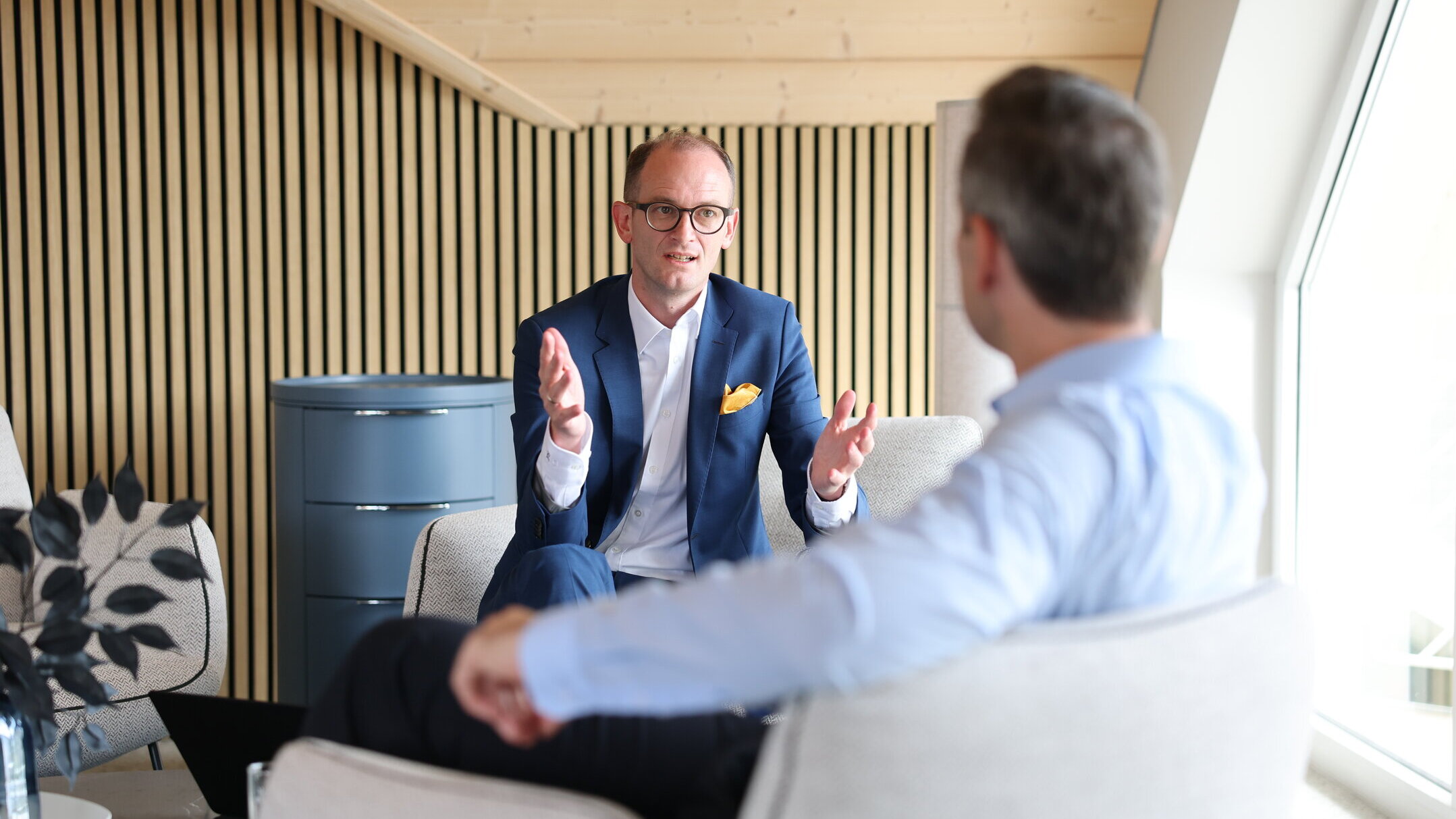 Beratungsgespräch bei Denkströme: Ein Manager im Anzug erklärt gestikulierend etwas in einem Lounge-Bereich, gegenüber sitzt ein Gesprächspartner.