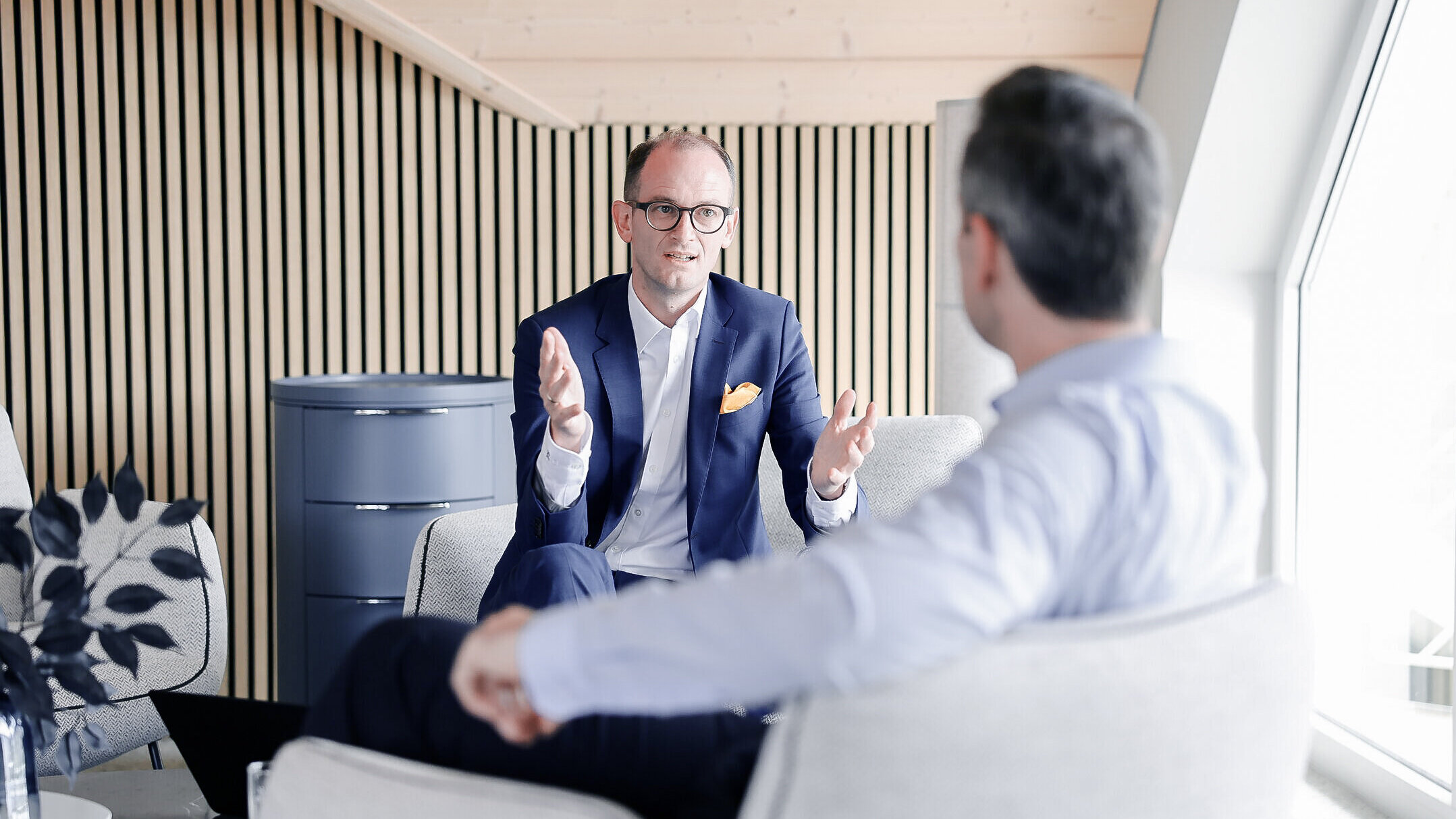 Beratungsgespräch bei Denkströme: Ein Manager im Anzug erklärt gestikulierend etwas in einem Lounge-Bereich, gegenüber sitzt ein Gesprächspartner.