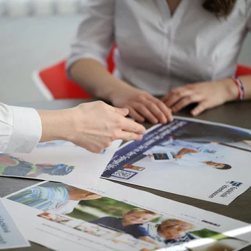 Drei Personen sitzen an einem Tisch und besprechen gedruckte Recruiting- und Employer-Branding-Materialien; eine Hand zeigt auf Layouts mit Fotos und Texten.