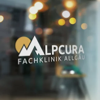Glasfront mit Logo-Fensterbeklebung der Alpcura Fachklinik Allgäu vor unscharfem Innenraum mit warmen Lichtreflexionen im Hintergrund.