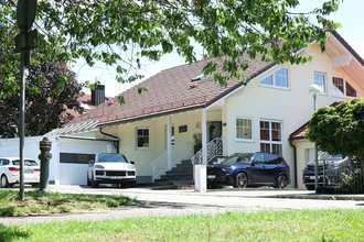 Außenansicht des Denkströme-Standorts in Kempten vom Haupteingang: helles Gebäude mit rotem Ziegeldach, Treppe zum Eingang, Denkströme-Branding an der Garage und geparkte Autos.
