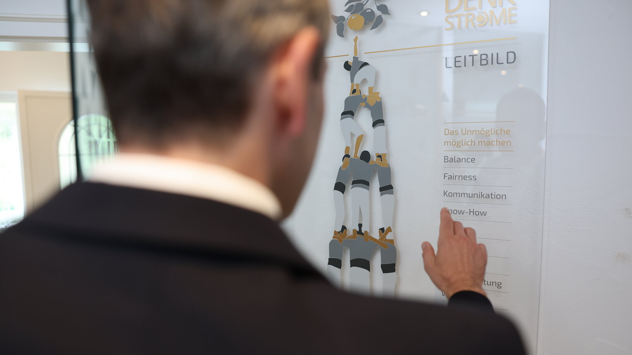 Mitarbeiter zeigt im Denkströme Büro auf das Leitbild an der Wand (Werte wie Balance, Fairness, Kommunikation und Know-how).