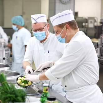 Küchenteam im DHZM – Mitarbeitende bereiten mit frischen Zutaten eine Mahlzeit in der Großküche zu, alle mit Hygienebekleidung und Mundschutz