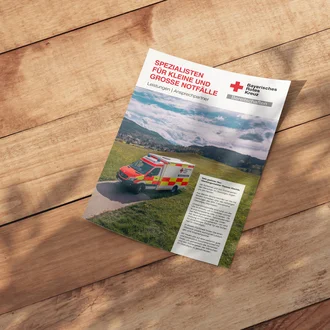 BRK-Factsheet des Bayerischen Roten Kreuzes auf Holztisch mit Rettungswagen vor Bergkulisse – Informationsbroschüre zu Bereitschaften und Notfallhilfe unter dem Motto ‚Spezialisten für kleine und große Notfälle‘