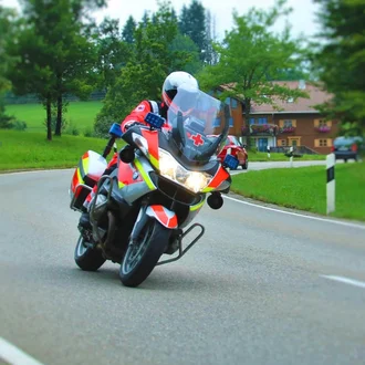 Einsatzfahrer des Bayerischen Roten Kreuzes auf schnellem Rettungsmotorrad in Kurvenfahrt durch ländliche Gegend – BRK-Motorradstaffel unterwegs zur Erstversorgung